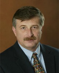 Kevin Campbell is an Alumni Board member who holds Emeritus status and he is wearing a black suit jacket and blue shirt with a tie and he has brown hair.