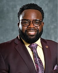 Alumni Board member Lester Booker Jr. in a very dark purple suit with a yellow shirt and pink swirly tie and he has short black hair and black glasses.