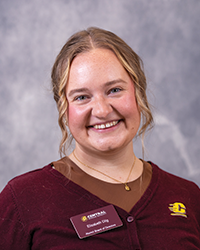 Lizzie is smiling for a headshot photo in front of a gray portrait backdrop. She is wearing a brown tank top with a maroon CMU cardigan, showing her CMU nametag and an embroidered action C.