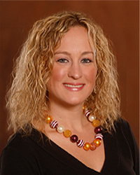 Michelle Rush is an Alumni Board member who holds Emeritus status and she is wearing a black top with a beaded necklace and has shoulder-length blonde wavy hair.