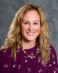 Morgan Hales is an Alumni Board member who holds Emeritus status and she is wearing a maroon shirt with pink flowers on it and she has long blonde hair.