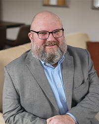 A headshot of a man with glasses and a beard, wearing a light blue collared dress shirt under a grey suit jacket.