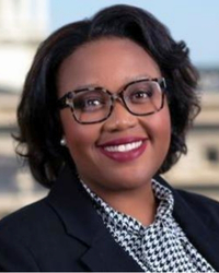 Headshot of Alumni Board of Directors member Karenia Randle standing in front of government buildings wearing a black blazer and black and white blouse and she has short black hair and glasses and is smiling with teeth.
