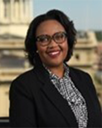 Headshot of Alumni Board of Directors member Karenia Randle standing in front of government buildings wearing a black blazer and black and white blouse and she has short black hair and glasses and is smiling with teeth.