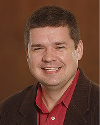 Ryan Fewins-Bliss is an Alumni Board member who holds Emeritus status and he is wearing a dark brown suit jacket and red shirt and has short brown hair.