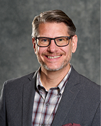 Scott Nadeau is an Alumni Board member and the current Vice President of the Alumni Board of Directors and he is wearing a gray suit with a plaid shirt and he has short hair and glasses.
