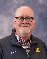 Alumni Board of Directors member Sean Hickey wearing a black athletic jacket with a plaid shirt and glasses in front of a gray background.