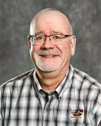 Alumni Board of Directors member Sean Hickey wearing a red and white plaid shirt and glasses in front of a gray background.