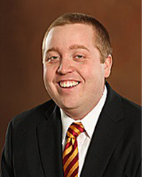 Spencer Long is an Alumni Board member who holds Emeritus status and he is wearing a black suit with a maroon and gold tie and has very short blonde hair.