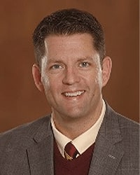Tom Olver is an Alumni Board member who holds Emeritus status and he is wearing a gray suit with a maroon vest and maroon and gold tie and he has short brown hair.
