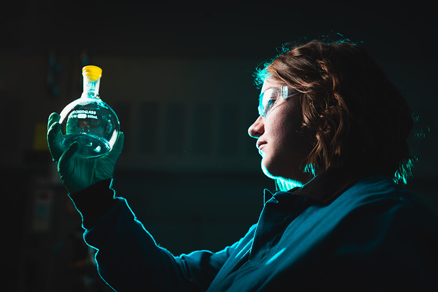 A female student looking at a boiling flask while wearing safety glasses and other lab safety equipment.