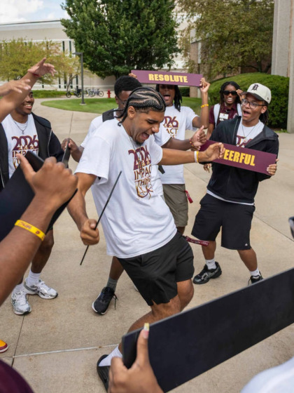 A group of CMU students wearing 2026 Impact shirts cheer and hold word signs as a student dances outdoors.