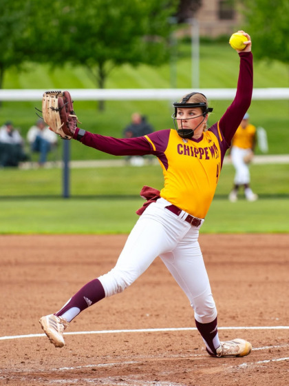 A CMU Chippewas softball pitcher wearing a face mask and a maroon and gold jersey winds up to deliver a pitch.