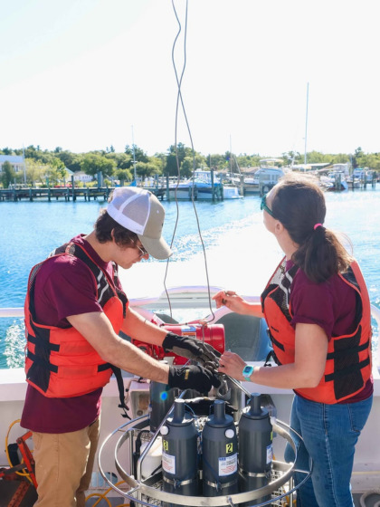 Two CMU student researchers wearing life jackets work with aquatic research equipment on a boat on open water.