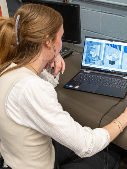 Over-the-shoulder view of a woman sitting at a desk, looking closely at a 3D CAD model displayed on a laptop.