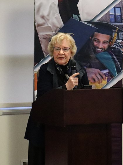 A woman in a black suit stands at a podium to the right of a College of Medicine banner.
