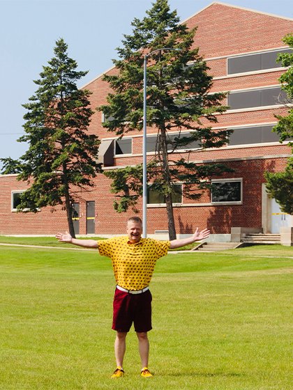 A man wearing a yellow polo and maroon shorts stands on grass in front of a building with his arms spread out.