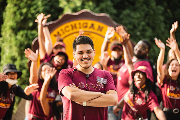 A group of students in Central Michigan University apparel standing and waving in front of the CMU seal.
