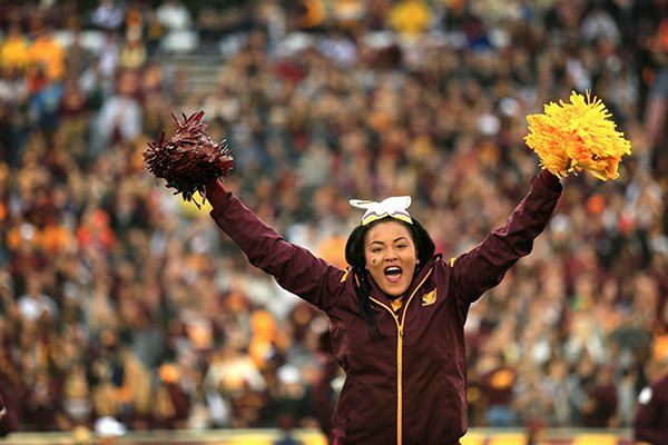 A cheerleader waving maroon and gold pom poms with a blurred crowd in the background.