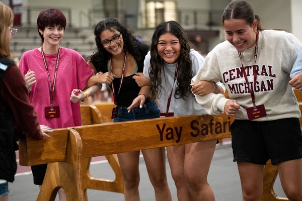 Four Central Michigan University students laugh and link arms behind a "Yay Safari" wooden beam during a CMU event.