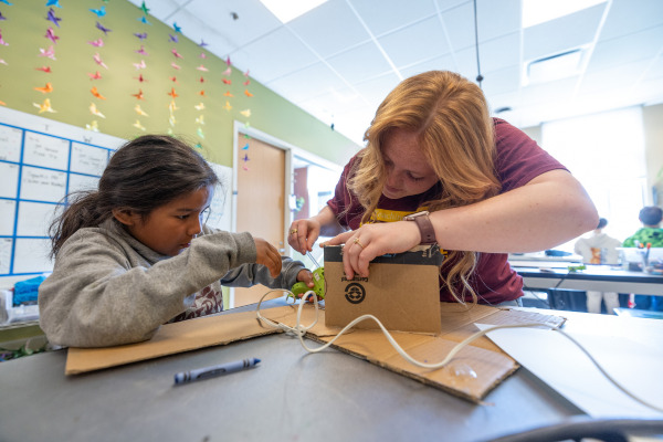 A female college student wearing a maroon t-shirt leans over a table to assist a young girl with assembling a cardboard structure using a hot glue gun.
