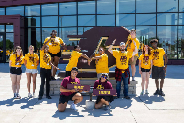 CMU students in maroon and gold IMPACT event shirts gather for a group photo in front of a large Action C sign.