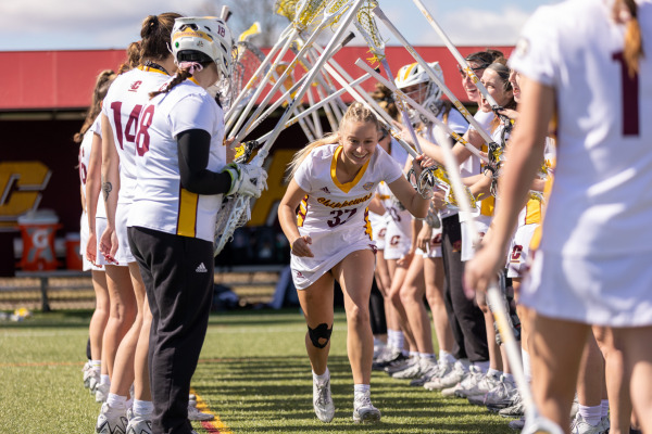 A smiling CMU women's lacrosse player in a white and gold jersey runs through a tunnel formed by teammates holding up their lacrosse sticks.