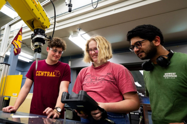 Three CMU students collaborate in an engineering lab while operating a yellow robotic arm.