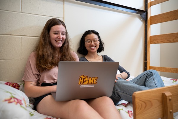Two Central Michigan University students, sitting on a bed against the wall, laugh together in a dorm room while looking at a laptop.