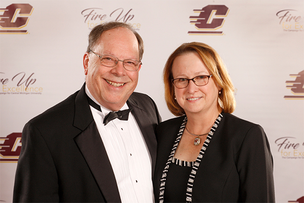 A man in a tuxedo smiles and poses next to a smiling woman wearing a black dress.