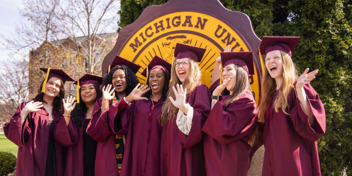 giving_fuelwhatmatters_700x350 Seven CMU graduates in maroon caps and gowns cheer and wave in front of the large Central Michigan University seal.