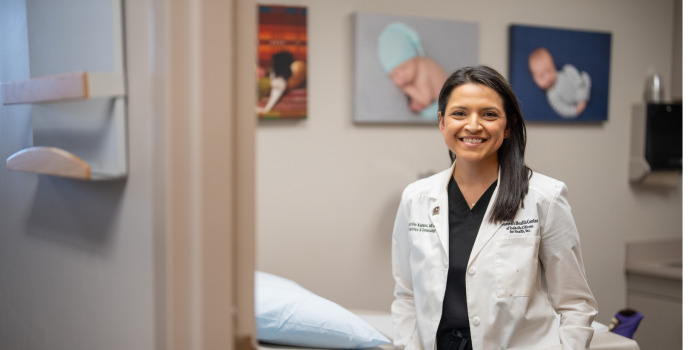 giving_initiatives_700x350 A female physician in a white lab coat over dark scrubs smiles inside a medical examination room with infant portraits on the wall behind her.