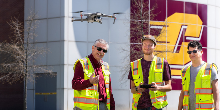 giving_transformational_700x350 Three men in high-visibility safety vests operate a drone outdoors in front of CMU's Engineering and Technology building, with a large Action C on the side of the building.