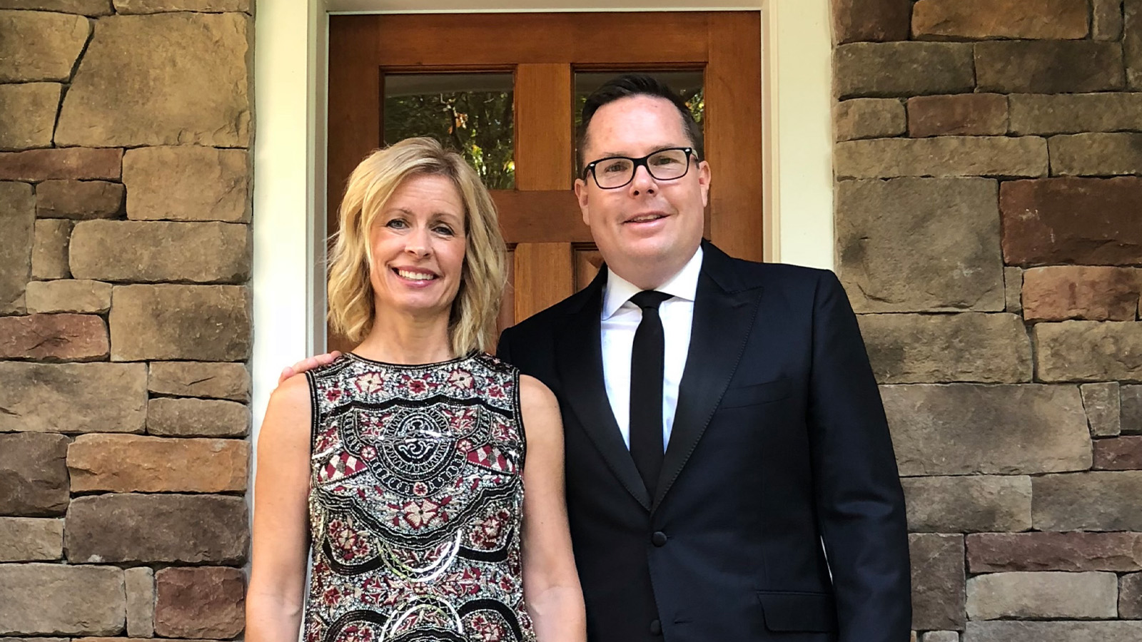 A man with glasses in a black suit, white dress shirt and black tie stands next to a woman in a black and red paisley dress. In the background is a door to a building.