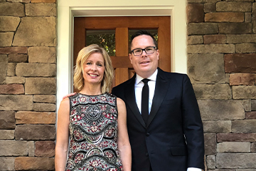 A man with glasses in a black suit, white dress shirt and black tie stands next to a woman in a black and red paisley dress. In the background is a door to a building.