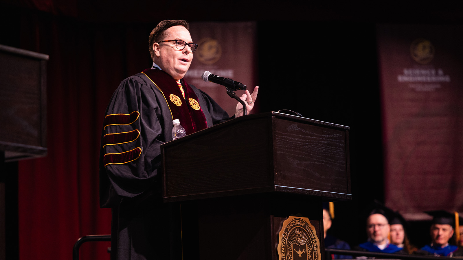 A man in a graduation gown is standing behind a podium and speaking into a microphone.