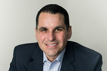 A man with dark hair wearing a navy blue suit jacket is smiling at the camera.