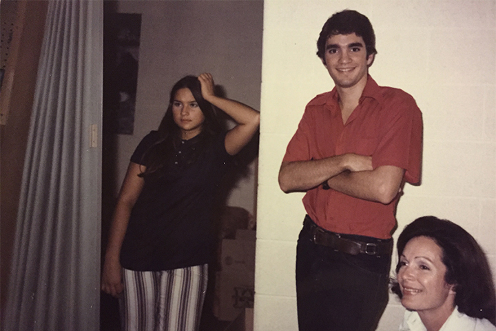 A man in a red shirt stands between two women. The woman on the left is wearing a black shirt and black and white striped pants. The woman on the right has a white shirt on. All three have dark hair.