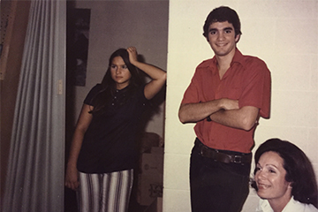 A man in a red shirt stands between two women. The woman on the left is wearing a black shirt and black and white striped pants. The woman on the right has a white shirt on. All three have dark hair.