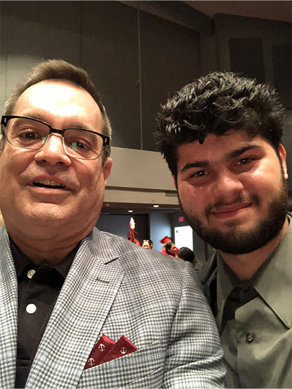 Selfie photo of a man wearing glasses and a checkered suit coat next to a man with dark hair and a beard wearing a green shirt.