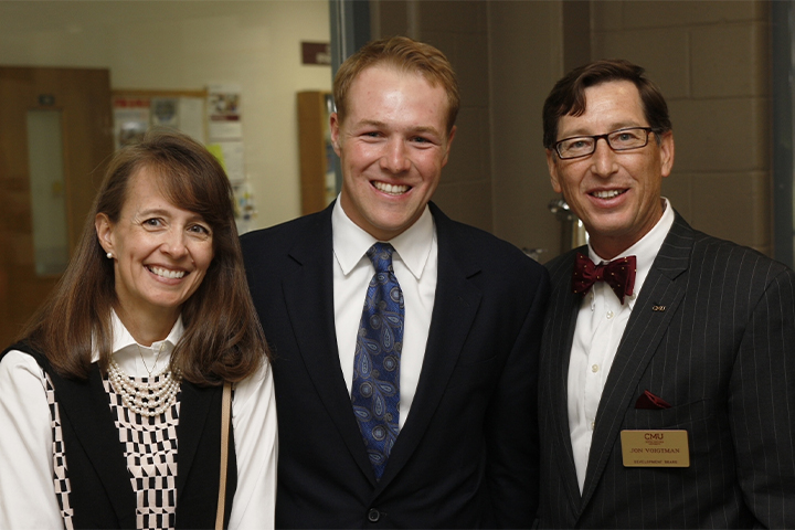 A younger man wearing a suit and tie stands between a middle aged woman in a sweater over a white dress shirt on the left and a middle aged man wearing glasses and a suit with a bowtie on the right.