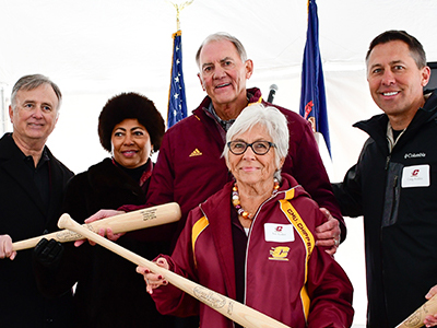 A woman in a maroon jacket stands with a group of people holding baseball bats.