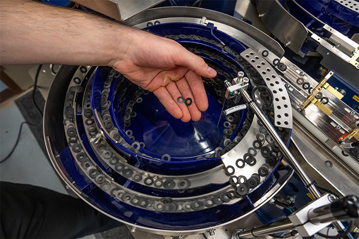 A hand holds two small circular nuts over a larger blue circular object with many circular spread out around it.
