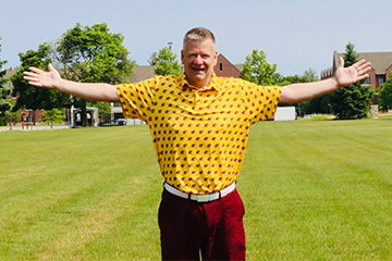A man wearing a yellow polo and maroon shorts stands with his arms spread out.