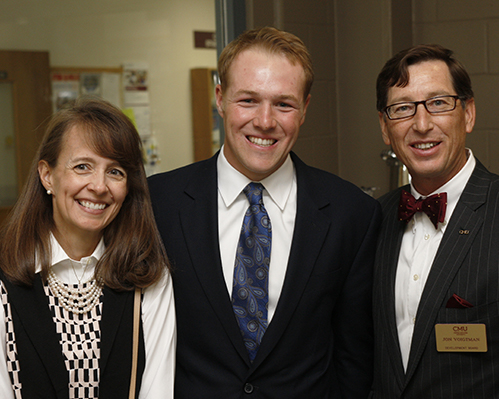 A man in a navy tie and suit coat stands with an alumni couple.