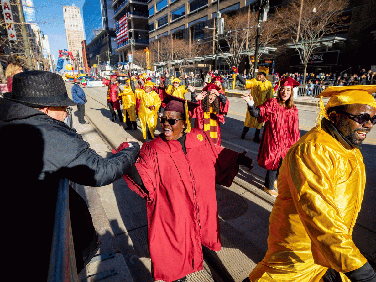 People wearing caps and gowns and marching in a parade. A woman is high fiving President Davies.