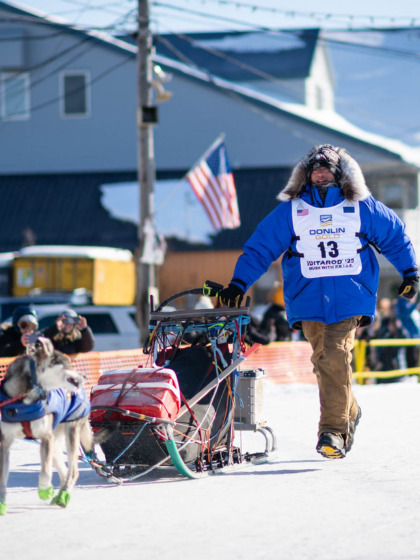 Keaton Loebrich, '22, wearing a blue parka, runs alongside his sled and dog team on a snow-covered street.