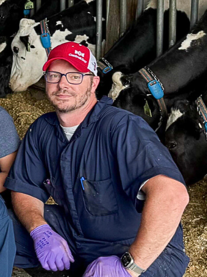 Casey Droscha, wearing a red and white baseball cap, glasses, and blue work coveralls, kneels in a barn with several cows visible in stalls behind him.