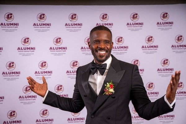 A man with a beard, wearing a black tuxedo with a bow tie, stands with open arms in front of a backdrop featuring the CMU Alumni logo.