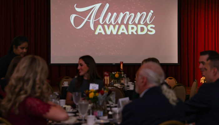 Attendees at an CMU Alumni Awards ceremony seated at round tables in a warmly lit room, with a projection screen displaying 'Alumni AWARDS' and maroon curtains in the background.